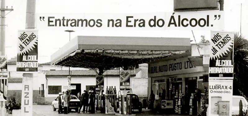 Imagem em preto e branco de um posto de gasolina com uma faixa escrito