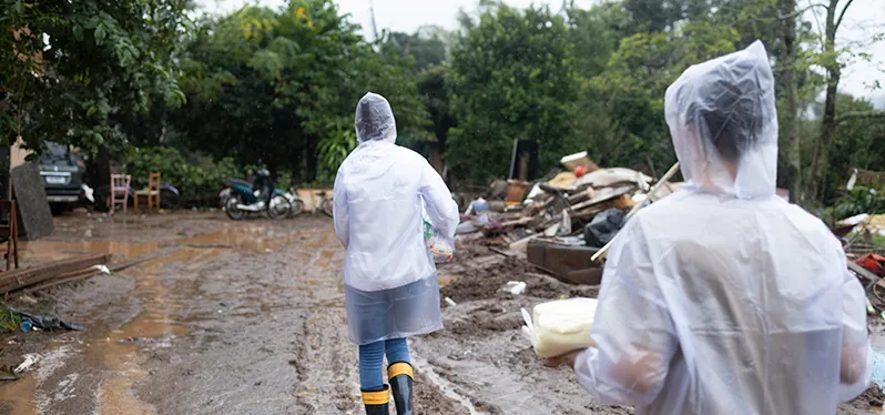 Na imagem, duas pessoas estão vistas de costas, ambas vestindo capas de chuva. Elas estão em um ambiente devastado, coberto por lama e resíduos sólidos, afetado por fortes temporais.