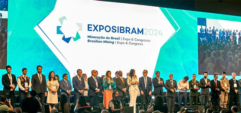 Na imagem, pessoas estão de pé no palco do evento, próximas a um painel de LED que exibe a inscrição