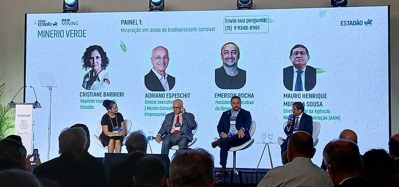 Painel de debate realizado em evento sobre mineração sustentável. Quatro participantes estão sentados no palco diante de uma plateia. Ao fundo, um telão exibe o tema “Mineração em áreas de biodiversidade sensível” e informações dos palestrantes.