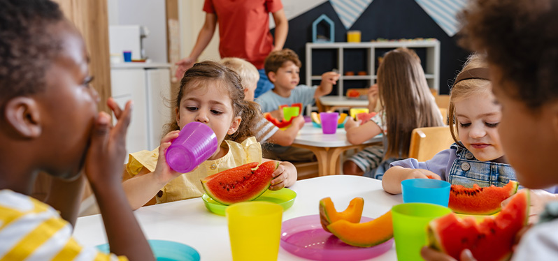 Crianças reunidas em mesas durante uma atividade coletiva em um ambiente de educação infantil, com acompanhamento de uma pessoa adulta ao fundo.