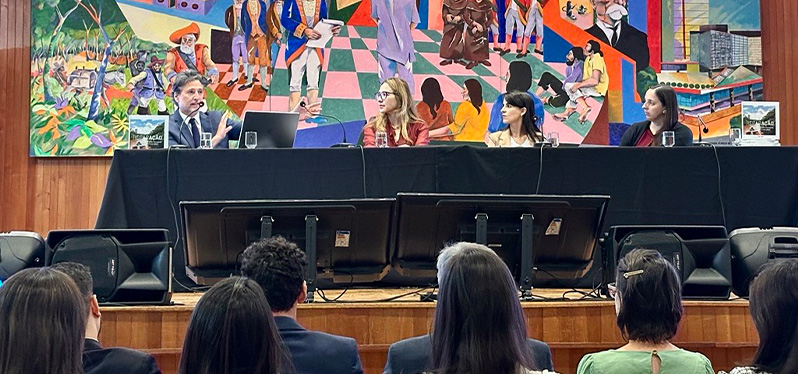 Mesa de debate do 1º Simpósio sobre Gestão de Desastres Socioambientais e Reparação em auditório. Quatro participantes estão sentados atrás de uma bancada com microfones, computadores e exemplares de publicações sobre o tema. À frente, o público acompanha a apresentação. Ao fundo, um grande painel artístico colorido cobre a parede do palco. A imagem registra um momento de discussão e troca de conhecimentos sobre gestão de desastres socioambientais e processos de reparação.